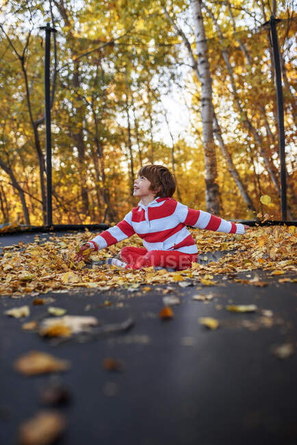 Niño sentado con las piernas cruzadas en un trampolín cubierto de hojas de otoño, Estados Unidos - foto de stock