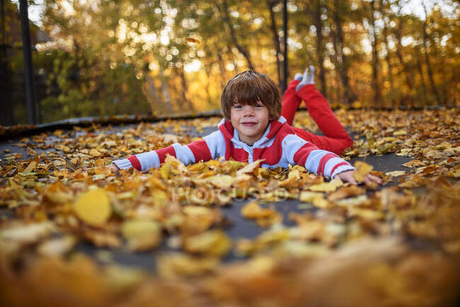 Портрет улыбающегося мальчика, лежащего на батуте, покрытом осенними листьями, США — стоковое фото