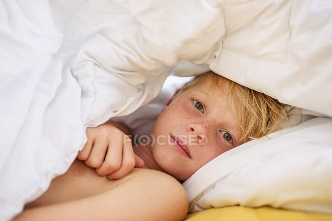Portrait of a boy lying in bed under a duvet — Stock Photo