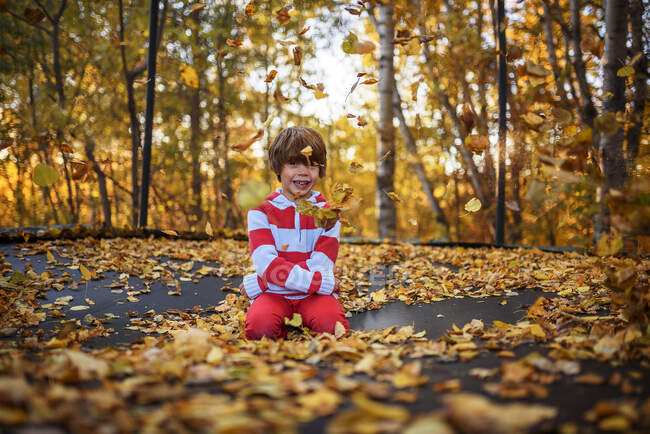 Smiling Boy sitting on a trampoline covered in autumn leaves, United States — Stock Photo