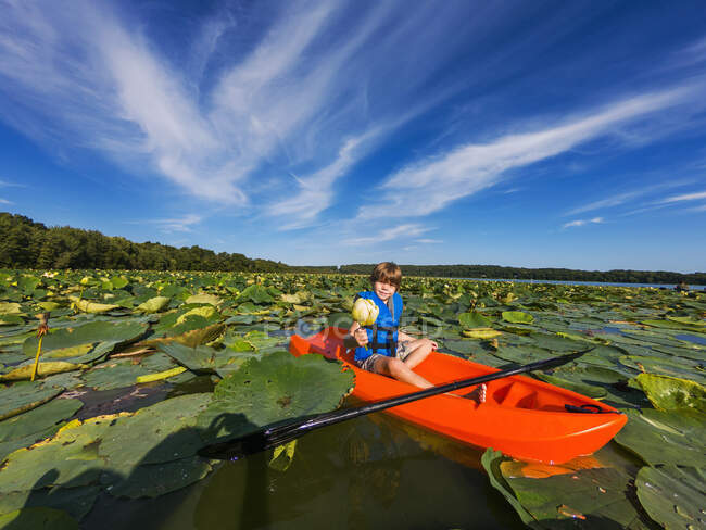 Niño sentado en un kayak sosteniendo una flor en un lago lleno de nenúfares, Estados Unidos - foto de stock