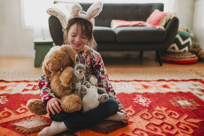 Menina sorridente vestindo orelhas de coelho sentado no chão com brinquedos macios — Fotografia de Stock