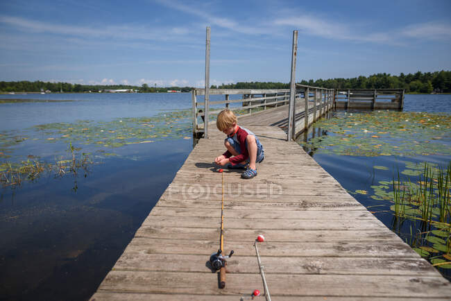 Niño sentado en un muelle adjuntando cebo a su caña de pescar, Estados Unidos - foto de stock
