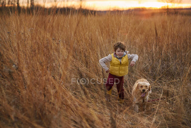 Garçon pourchassant son chien à travers un champ au coucher du soleil, États-Unis — Photo de stock