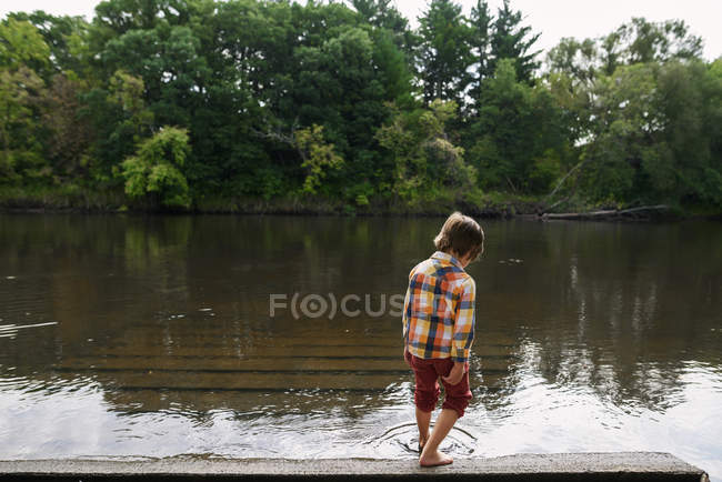 Niño parado junto a un río sumergiendo su dedo del pie en el agua, Estados Unidos - foto de stock
