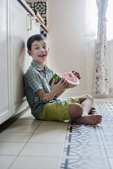 Усміхнений хлопчик сидить на підлозі на кухні, тримаючи шматочок кавуна — стокове фото