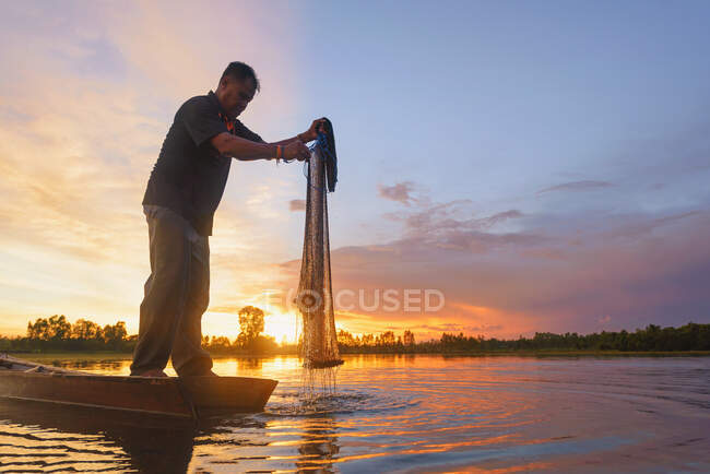 Silueta de un pescador parado en un barco sosteniendo una red de pesca al atardecer, Tailandia - foto de stock