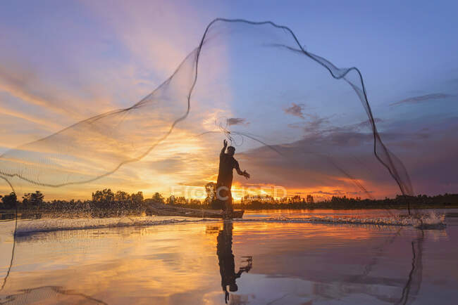 Silhouette d'un pêcheur debout dans un bateau jetant un filet de pêche au coucher du soleil, Thaïlande — Photo de stock