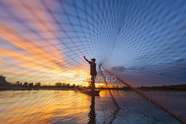 Silueta de un pescador de pie en un barco que lanza una red de pesca al atardecer, Tailandia - foto de stock