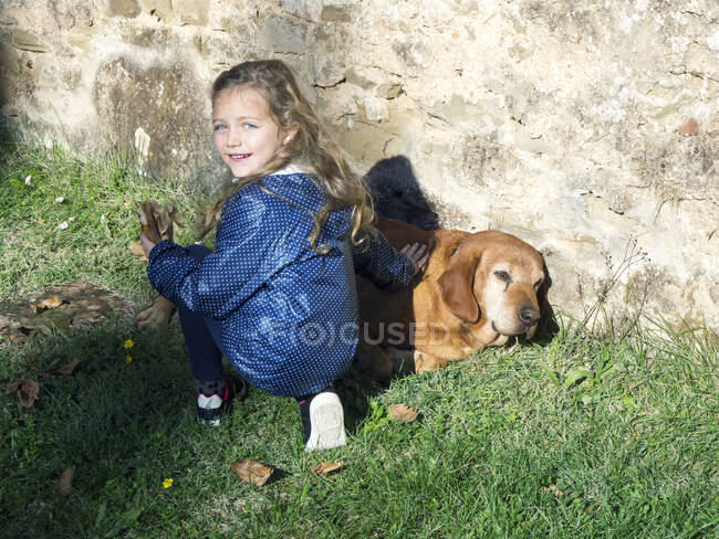 Chica sonriente agachada por una pared acariciando a un perro, Italia - foto de stock