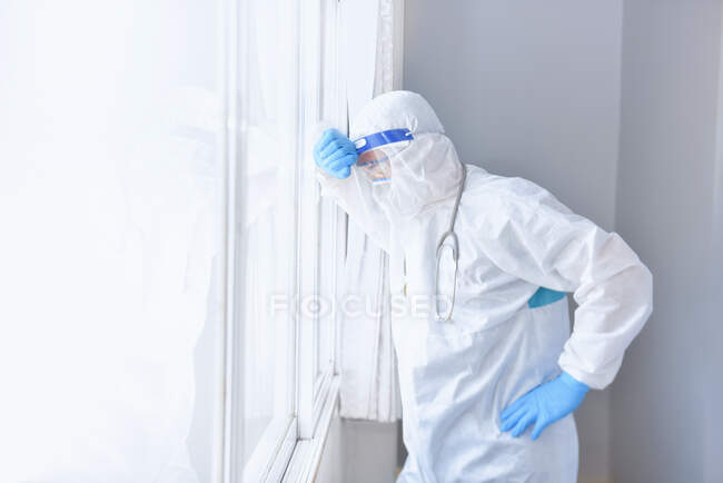 Tired doctor wearing PPE leaning against a window, Thailand — Stock Photo