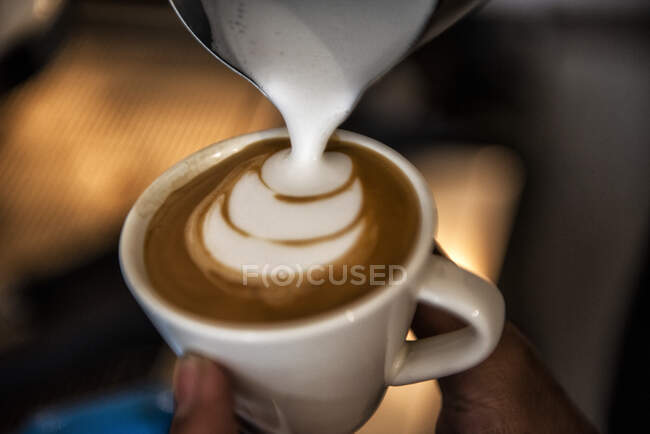 Primer plano de una persona que vierte microespuma en una bebida de café con leche piccolo para hacer un patrón decorativo - foto de stock