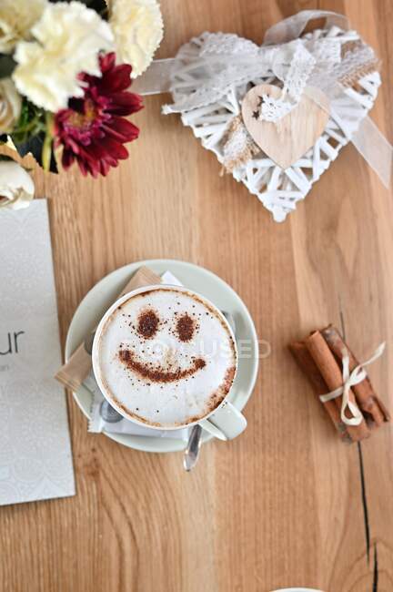 Cup of coffee with a smiley face froth decoration next to a vase and heart shape decoration — Stock Photo