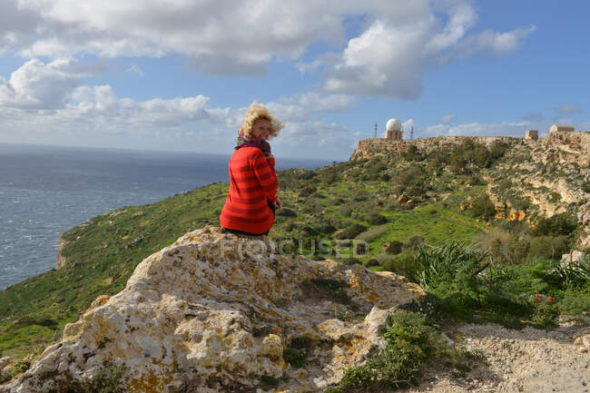Rückansicht einer Frau, die auf einem Felsen sitzt und auf die Dingli-Klippen blickt, mit Dingli-Radarstation in der Ferne, Malta — Stockfoto