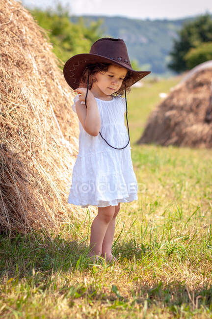 Menina vestindo um chapéu de cowboy em pé em um campo por um fardo de feno, Bulgária — Fotografia de Stock