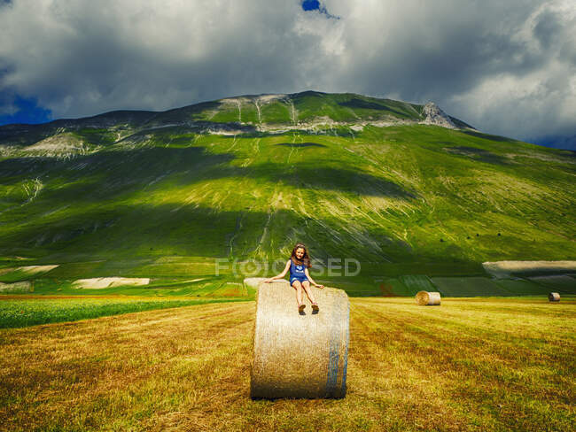 Chica sentada en una bala de heno en un campo, Castelluccio di Norcia, Umbría, Italia - foto de stock