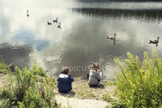 Vista trasera de dos chicas sentadas junto a un lago mirando cisnes, Polonia - foto de stock