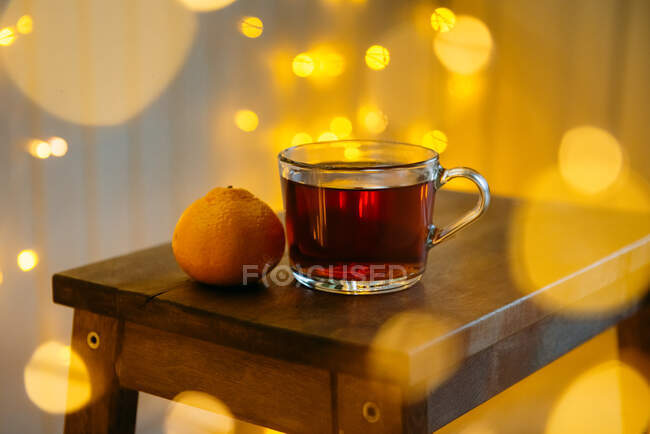 Xícara de chá e uma tangerina em uma mesa com decorações de luz de fada — Fotografia de Stock
