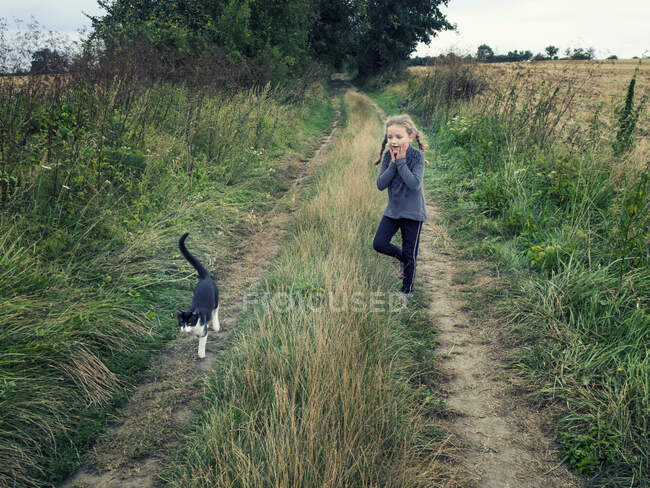 Chica de pie en un camino rural mirando a un gato, Polonia - foto de stock