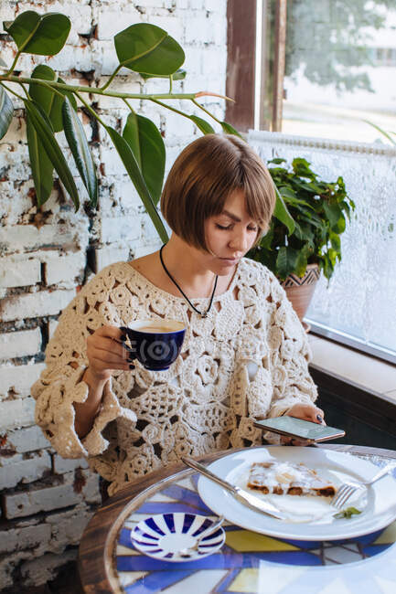 Mujer sentada en un café bebiendo café y mirando su teléfono móvil - foto de stock