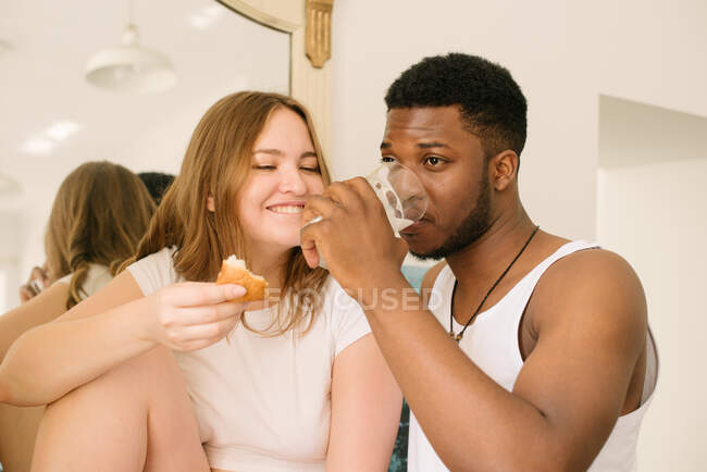 Casal de raça mista comer um croissant com geléia e beber um copo de leite — Fotografia de Stock