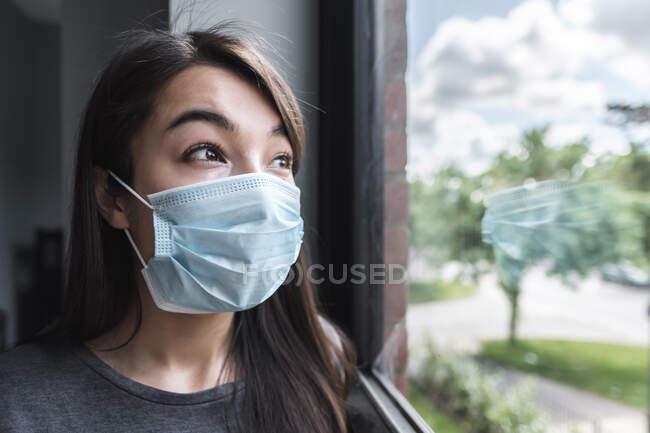 Woman wearing a face mask looking out of a window during lockdown - foto de stock