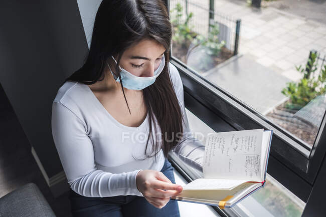 Женщина в маске сидит у окна и читает во время изоляции — стоковое фото