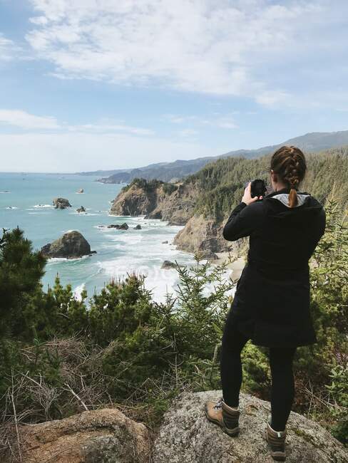 Rear view of a woman standing on cliffs taking a photo, Samuel H Boardman State Park, Oregon, USA — Stock Photo