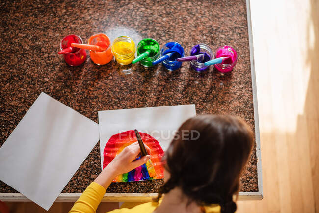 Chica sentada en la cocina pintando un arco iris - foto de stock