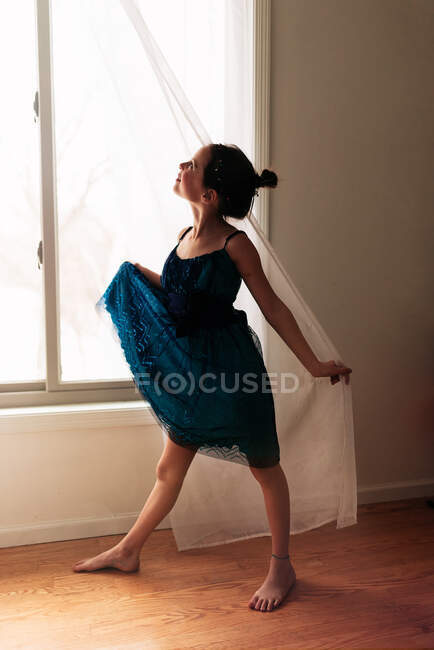 Portrait of a young girl standing by a window holding the curtain — Stock Photo