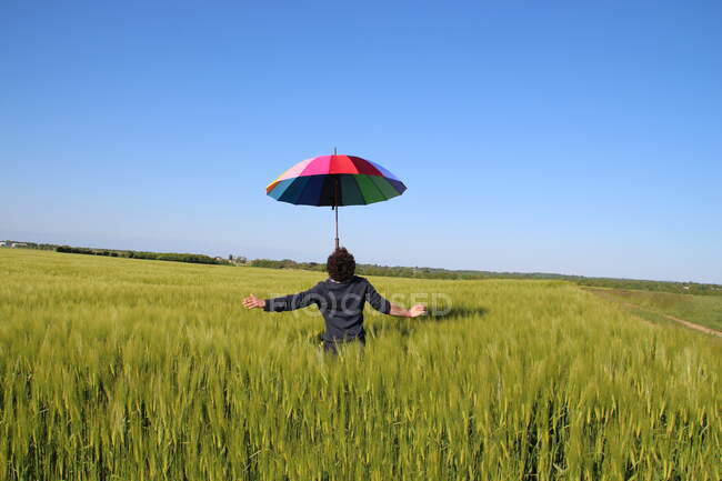 Homme debout dans un champ de lin équilibrant un parapluie multicolore sur son visage, France — Photo de stock