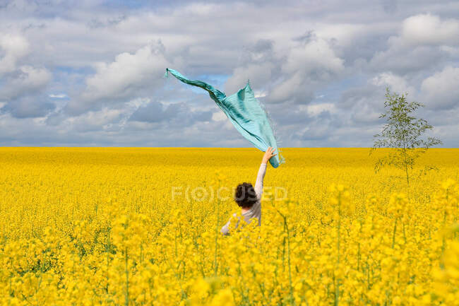 Mujer de pie en un campo de colza sosteniendo su bufanda en el aire, Francia - foto de stock