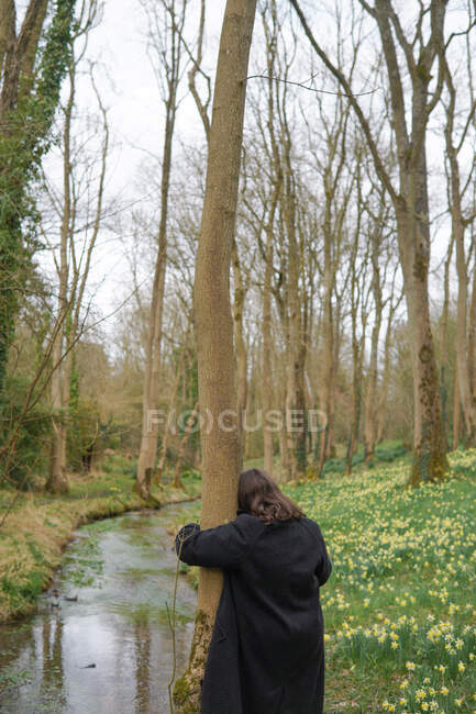 Rear view of a woman standing by a river hugging a tree, England, UK — Stock Photo