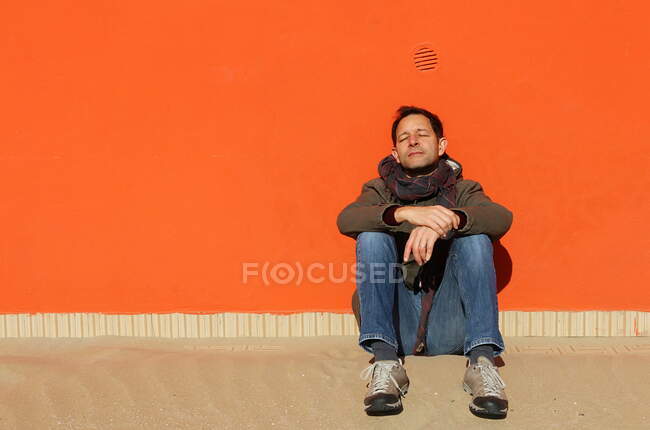 Retrato del hombre sentado en la playa apoyado contra una pared naranja al sol, Rimini, Emilia-Romaña, Italia - foto de stock