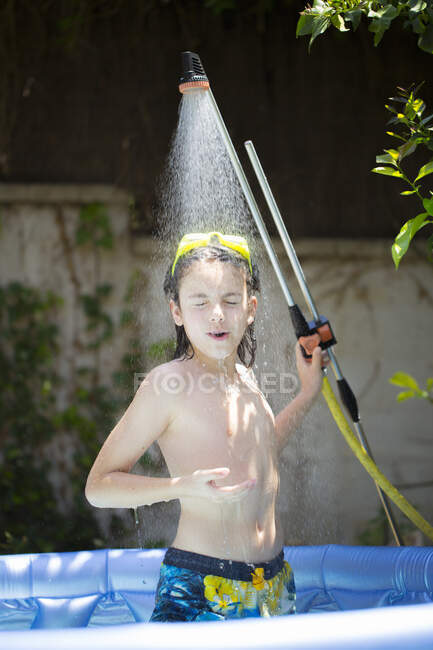 Boy sitting in a paddling pool cooling off under a shower — Stock Photo