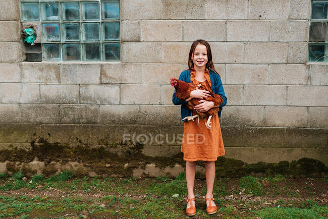 Chica sonriente de pie en una granja sosteniendo un pollo, EE.UU. - foto de stock