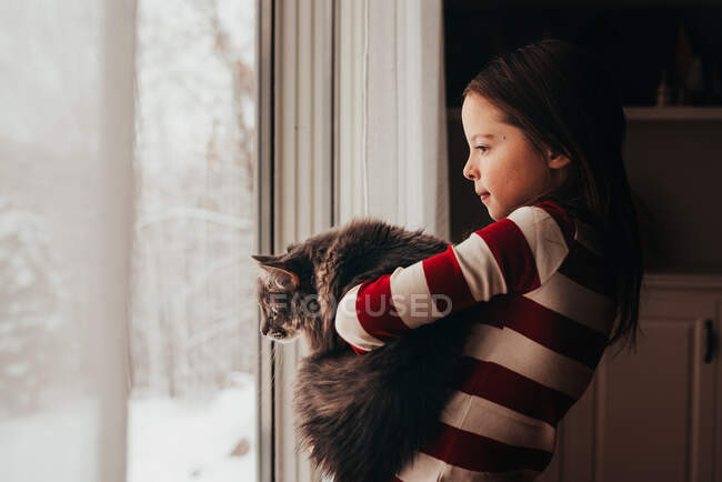 Menina em pé por uma janela abraçando seu gato — Fotografia de Stock