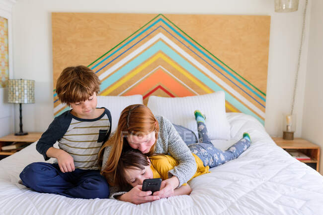 Мать и дети лежат вместе на кровати и смотрят на смартфон — стоковое фото