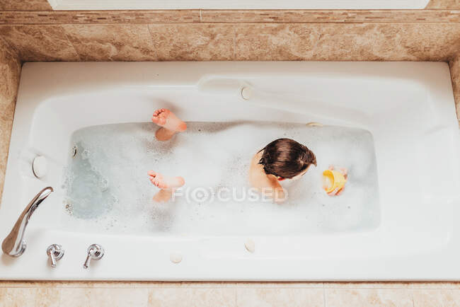 Vista aérea de un niño acostado en su frente en un baño de burbujas jugando con una taza de plástico - foto de stock