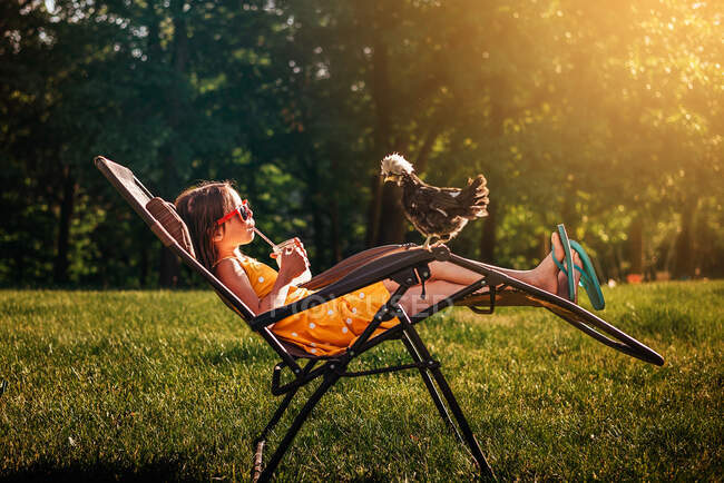 Happy girl sitting in the garden with a chicken on her chair — Stock Photo
