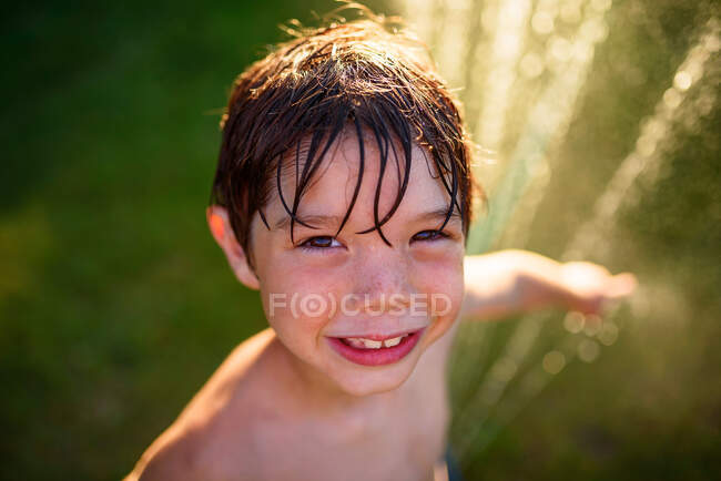 Ritratto di un ragazzo sorridente in piedi accanto a un irrigatore d'acqua in giardino, USA — Foto stock