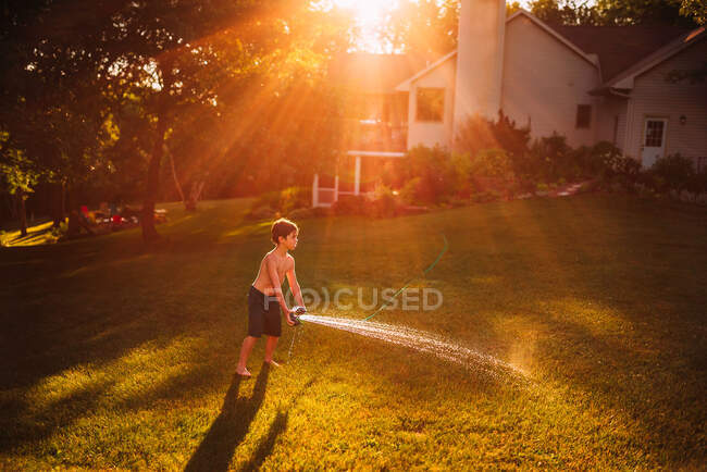 Niño de pie en un jardín jugando con un rociador de agua, EE.UU. - foto de stock
