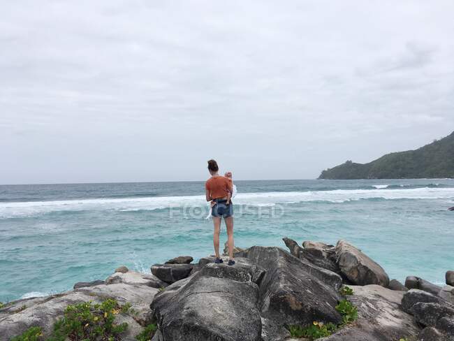 Donna in piedi sulle rocce in riva al mare con il suo bambino, Seychelles — Foto stock