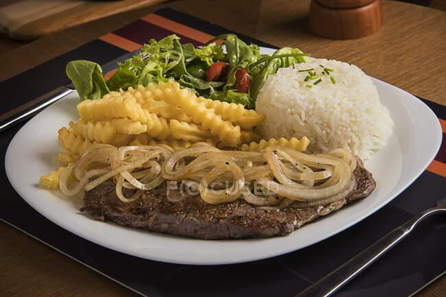 Bistecca con cipolle fritte, riso, patatine fritte e insalata — Foto stock