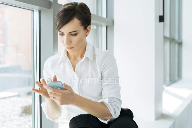 Mujer de negocios sentada en un alféizar de la ventana usando su teléfono móvil - foto de stock