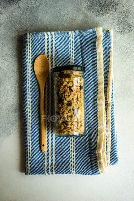 Glass jar filled with granola next to a wooden spoon on a tea towel — Stock Photo