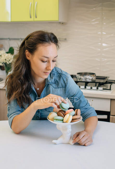 Woman sitting in a kitchen choosing a macaroon — Stock Photo