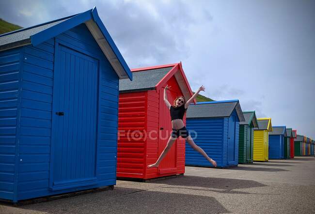 Chica saltando en el aire por cabañas de playa, Whitby, Yorkshire, Inglaterra, Reino Unido - foto de stock