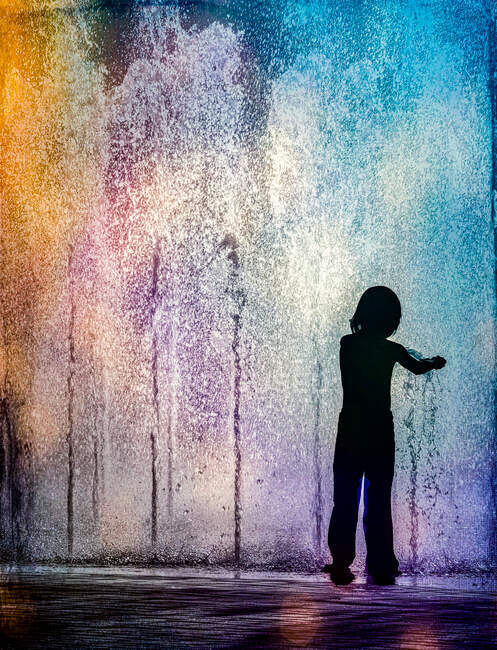 Silhouette of a child standing in a multi coloured water fountain, USA — Stock Photo