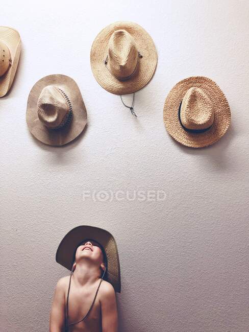 Garoto sorridente usando um chapéu de palha e olhando para chapéus de palha pendurados em uma parede — Fotografia de Stock